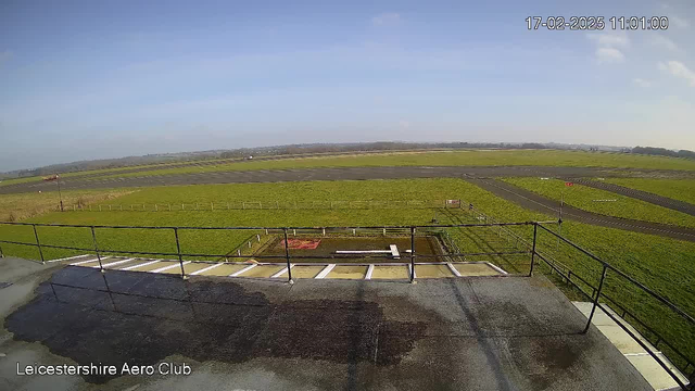 A clear view of a grass airfield under a bright blue sky. In the foreground, there is a railing and a flat surface with some water puddles. The airfield is divided by visible runway markings and has a few scattered objects like signs and fences. In the distance, low hills can be seen against the horizon. The overall scene conveys a sense of open space and tranquility.