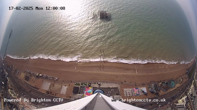 Aerial view of a beach with waves gently lapping the shore. The beach is sandy and stretches horizontally across the bottom of the image. A group of people can be seen walking near the water's edge. In the distance, a ruined pier juts out into the ocean. To the right, there is a promenade with colorful structures and a circular pool. The sky is light and the ocean water is a mix of green and blue, reflecting sunlight. The date and time are displayed in the top left corner.