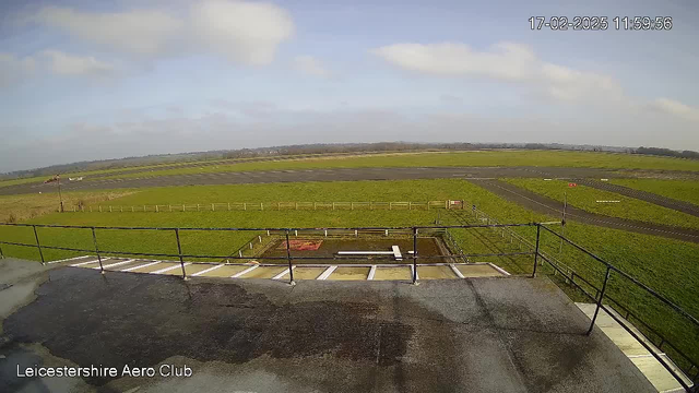 A view from a webcam overlooking a green, grassy field and an airstrip at the Leicestershire Aero Club. The foreground includes a flat rooftop surface with a railing. In the distance, there are scattered clouds in a blue sky, and an airplane is visible on the runway. The landscape is bordered by a fence and stretches towards the horizon, where more land can be seen.