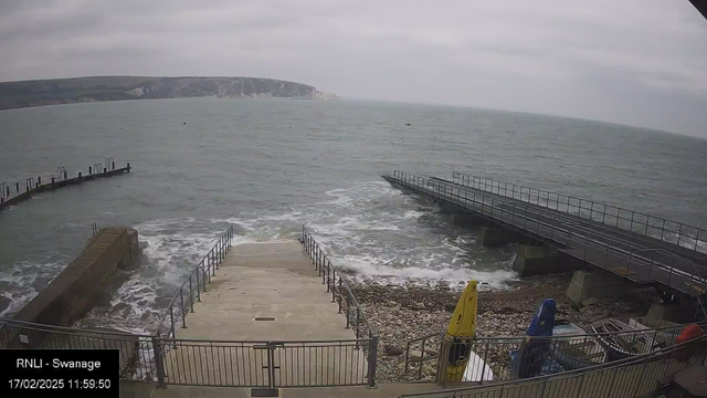 A cloudy seaside scene shows gentle waves lapping at a rocky shore. A set of concrete steps leads down from a raised area to the water. To the left, a small pier extends out over the sea, while another broader dock is visible to the right. Several kayaks in bright colors, including yellow and blue, are stacked near the edge. The coastline features green hills in the distance, with faint white cliffs visible under the overcast sky. The image captures a tranquil, coastal atmosphere.