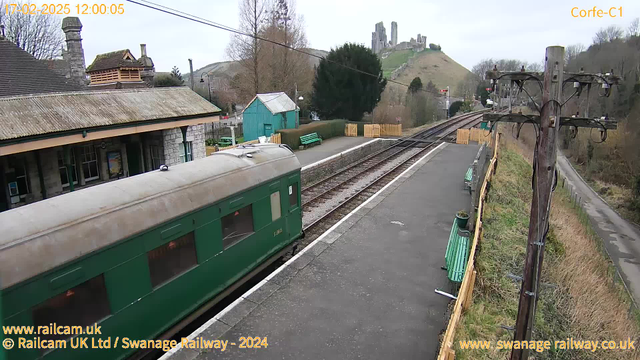 A view of a train station with a green train parked on the platform. The platform is lined with wooden benches and features a small wooden fence. In the background, a hilly landscape is visible with a castle on a hilltop. There are trees surrounding the station, and a few buildings are seen, including a turquoise shed. The sky is overcast, contributing to a gray atmosphere. The railway tracks extend away from the viewer.