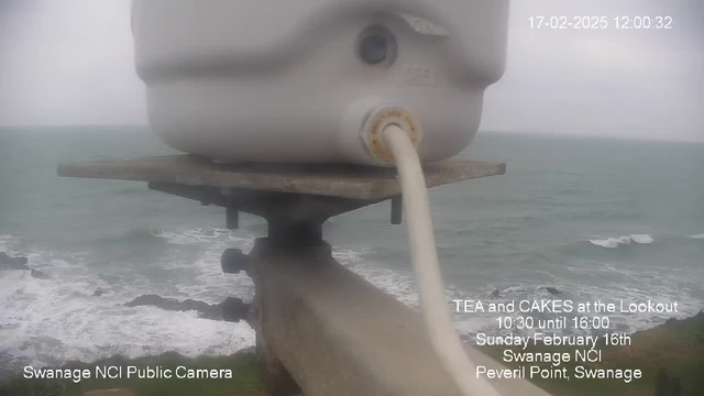 A close-up view of a white container on a metal platform, which is situated above a rocky coastline. A hose is attached to the container, leading downward. The ocean is visible below with gentle waves, and the sky appears overcast, suggesting a cloudy day. Text indicating the time and date is present in the top right corner, as well as information about a tea and cake event happening at a lookout point nearby.