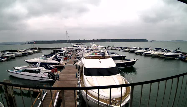 A harbor scene on a cloudy day with various boats and yachts docked in the water. The foreground features a prominent white yacht with a black canopy. There are several other boats of different sizes and colors, including some blue and striped boats. The water appears calm, and the shoreline can be seen in the distance, with greenery along the edge. A wooden dock is visible, leading to the boats, and the overall atmosphere is serene and tranquil.