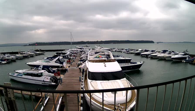 A view of a marina on a cloudy day, featuring numerous boats and yachts moored in calm water. The image shows a wooden dock leading to several vessels, including a large white yacht in the foreground. In the background, more boats are visible, along with a shoreline partially obscured by clouds.