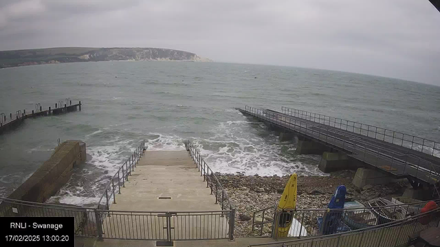 A coastal scene featuring a grey sky with clouds. The ocean is visible, with gentle waves lapping against the shore. There are steps leading down to the water, bordered by a metal railing. To the left, a small dock extends into the water with black posts. On the right, another wooden pier juts out over the sea. Various boats, including one yellow kayak and one blue kayak, are resting on the ground near the water's edge, surrounded by rocky and gravelly terrain.