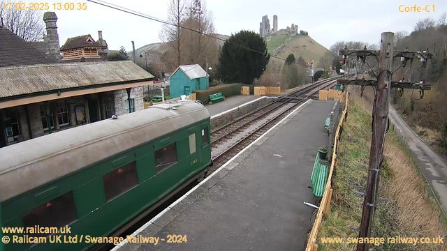 A green train car is visible on the left side of the image, parked at a railway station platform made of gray stone. In the background, a hill features a castle ruins at the top. There are several green benches along the platform, and a wooden fence separates the platform from a grassy area. A wooden utility pole with wires is positioned on the right side of the image. The sky is overcast, creating a muted lighting atmosphere.