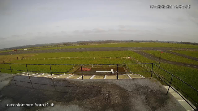 A wide shot of an aerodrome on a cloudy day. The foreground shows a railing, with a slightly wet surface, leading to a grassy area. The middle ground features a large, open runway, partially covered in asphalt with white markings. There is a patch of grass on one side and a red sign in the distance. The background consists of a cloudy sky, with hills visible in the far distance. The timestamp at the top indicates it is February 17, 2025, at 13:00.