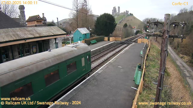 A green railway carriage is shown in the foreground, partially obscured by the camera's angle. The background features a train station platform with wooden benches and a small blue building. In the distance, there is a hill topped with ruins, likely a castle. Trees are visible around the station, and there is a telephone pole with wires on the right side of the image. The scene appears to be bright and clear, suggesting daytime conditions.