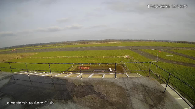 A view from a rooftop over an airfield. The foreground features a railing and a dark surface below it. The airfield has green grass and several runways marked in black. In the distance, a small red aircraft windsock is visible. The sky is mostly cloudy with some light grey clouds, and the landscape includes a flat horizon with patches of trees. The image shows a typical day at a small aerodrome.