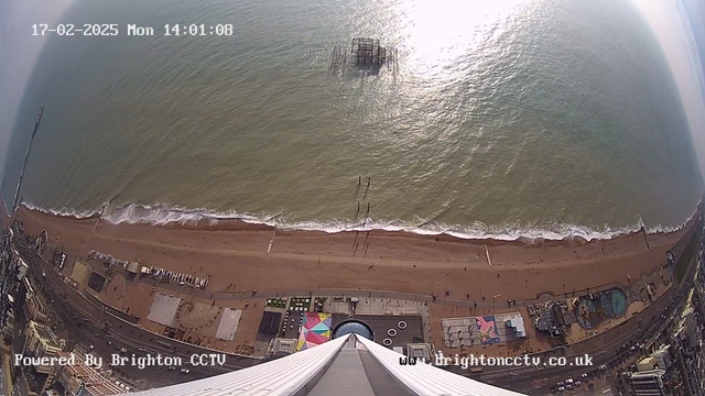 Aerial view of a sandy beach with gentle waves lapping at the shore. In the distance, there is a partially submerged pier structure. The beachfront is lined with various buildings and amusement rides, some of which have bright colors. Shadows are cast onto the sand from people walking along the beach. Above the scene, a clear sky meets the water, which is reflecting sunlight. A timestamp reads "17-02-2025 Mon 14:01:08" in the upper left corner, with a "Powered By Brighton CCTV" label at the bottom.