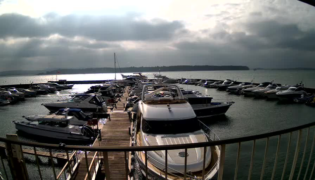 A view of a marina with several boats and yachts docked. In the foreground, a large white motorboat is moored beside a wooden dock, which leads to other boats. The water is calm with occasional ripples, and there are cloudy skies overhead, filtering the light. In the background, additional boats are lined up along the marina, with a shoreline visible in the distance.