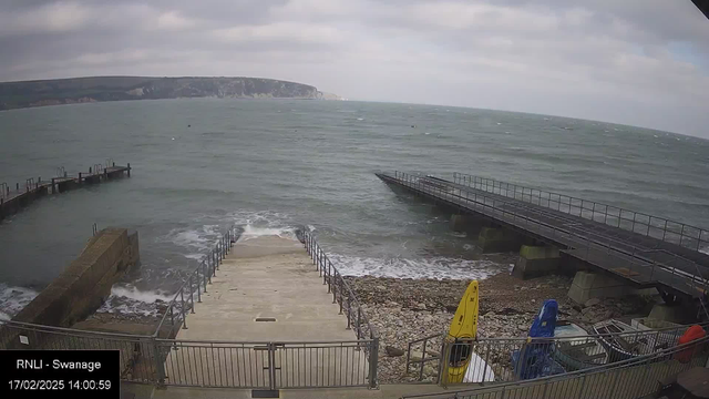 A seaside view featuring a calm, grayish sea under overcast skies. In the foreground, a set of stairs leads down to the water, flanked by a rocky shoreline. To the right, several boats in bright colors, including yellow and blue, are secured next to a wooden pier. The pier extends into the water, with a railing visible, and the shoreline in the background includes cliffs that rise gently.