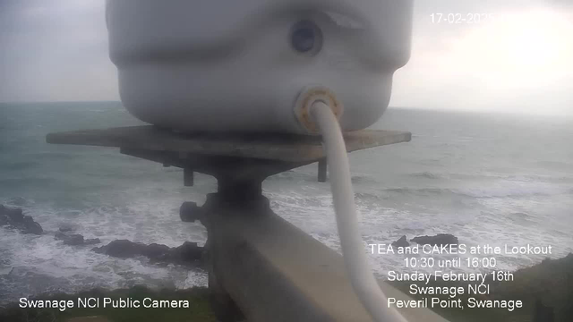 A view from a webcam overlooking a choppy sea with waves crashing against rocky formations at the shore. The sky is overcast and gray, indicating a cloudy day. In the foreground, a white cylindrical object is mounted on a horizontal platform, possibly part of the webcam apparatus. Text overlay at the bottom left reads "Swanage NCI Public Camera," and additional text provides details about an event featuring tea and cakes at the Lookout on February 16th, 2020.