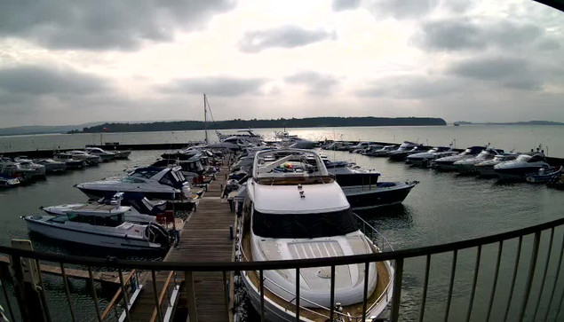 A view of a marina filled with various boats, including larger yachts and smaller vessels, docked at wooden piers. The water is calm with a few ripples, reflecting the overcast sky, which has thick clouds. In the background, there are green hills lining the shore, and the scene conveys a peaceful, nautical atmosphere.