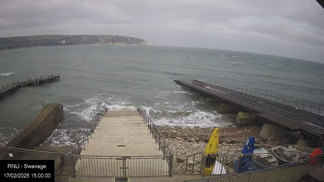 A view of a seaside area featuring a set of stone steps descending to the water. On the left, there is a concrete structure with a railing, and several wooden docks extend into the water. The sea is choppy and gray under overcast skies, with some white caps visible on the waves. In the foreground, there are a couple of colorful kayaks—one yellow and one blue—positioned on the rocky shore. The scene conveys a sense of a cool, breezy day by the coast.