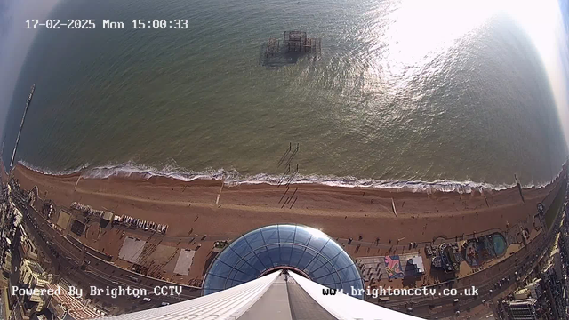 Aerial view of a beach and ocean, showing a sandy shoreline with gentle waves lapping at the shore. An old pier structure is partially submerged in the water. Below, there are beachside attractions and buildings, including amusement rides. The camera captures a clear day with sunlight reflecting off the water. The date and time are displayed in the corner.