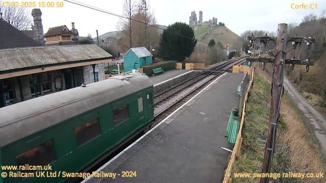 A green train carriage is parked at a railway station platform with a stone building in the background. In the distance, castle ruins are visible on a hilltop. There are green benches along the platform and a wooden fence nearby. Power lines and poles are present on the right side of the image. The weather appears overcast, with a gray sky.