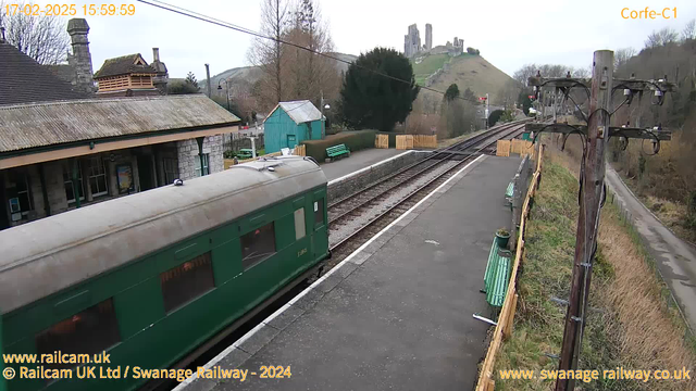 A green train car is seen in the foreground, partially on the platform of a railway station. The station has a stone building with a sloped roof, and a bright blue shed in the background. There are several green benches along the platform and a wooden fence separating the area from a road on the right side. In the distance, there are hills with a castle-like structure atop one, visible under an overcast sky. Power lines and a utility pole are located on the right side of the image.