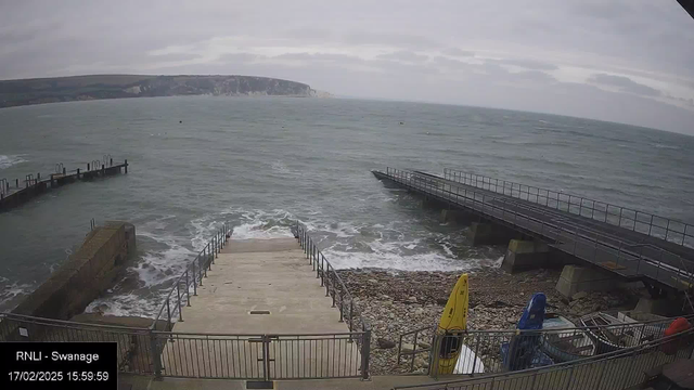 A cloudy view of a seaside area. In the foreground, there are steps leading down to the water, flanked by a railing. To the left, a small pier extends into the sea, and to the right, another pier reaches out further, both supported by large concrete structures. The water is choppy with visible waves, and some rocks are scattered along the shore. A few brightly colored kayaks are seen at the edge of the image, next to the railing. The landscape in the background includes hills that rise up towards the horizon.