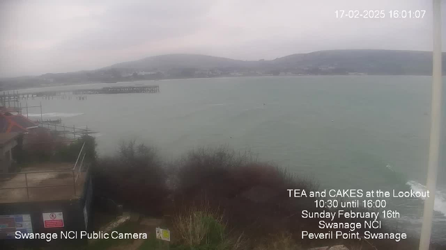 The image shows a cloudy coastal scene with a view of a body of water. In the foreground, there are buildings partially obscured by shrubs and grass. To the left, scaffolding is visible near the edge of the structure. The water appears calm with small waves, and a distant wooden pier extends into the water. The background features hills rising from the shoreline and a gray sky, indicating overcast weather. Text overlays indicate information about a local event at Swanage NCI.