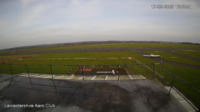 A wide-angle view of an airport runway taken from an elevated position. The foreground features a flat rooftop with metal railings. In the background, there is a runway extending into the distance, bordered by green grass and a wooden fence. A small airplane is visible on the runway, positioned toward the bottom right. The sky is overcast with light clouds, and the ground is wet, suggesting recent rain. The runway has markings and a windsock on the left side, indicating wind direction.