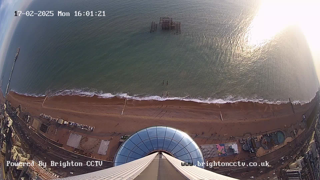 Aerial view overlooking a beach and sea. The camera is positioned high above, showing a curved rooftop structure below. The sandy beach stretches out with gentle waves lapping at the shore. A pier is visible extending into the water, and some people can be seen walking along the beach. The image is taken during the late afternoon, with sunlight shimmering on the water's surface. The sky is mostly clear with a hint of clouds.