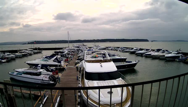 A marina scene with numerous boats docked in a calm waterway. Several white and blue yachts are visible, arranged closely together. A wooden dock is present in the foreground. The sky appears overcast, with gray clouds and a soft light, indicating early morning or late afternoon. In the background, gently rolling hills are outlined against the horizon, with some trees partially visible.