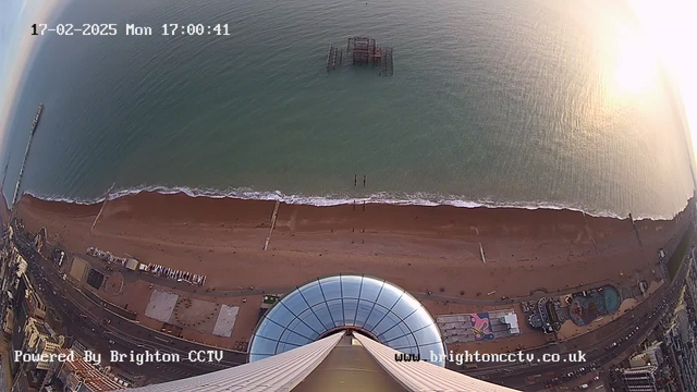 Aerial view of a beach at sunset, showcasing a sandy shore meeting gentle waves. In the water, a partially submerged pier is visible. The foreground features a circular structure with a reflective surface, likely part of a tower or viewing platform. The beach is lined with a few structures, including amusement rides and beachside facilities. The sky transitions from blue to soft hues of orange and pink, illuminated by the setting sun.