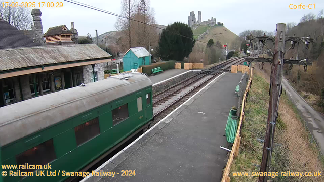 A green train carriage is partially visible on the left side of the image, sitting on a platform. The platform features green benches and a stone wall. In the background, a charming stone building with a slate roof can be seen, along with a small, teal-colored shed. There are trees surrounding the area, and a hill with ruins of a castle is visible in the distance. The sky is overcast. Power lines and a wooden pole with electrical wires are located on the right side of the image.