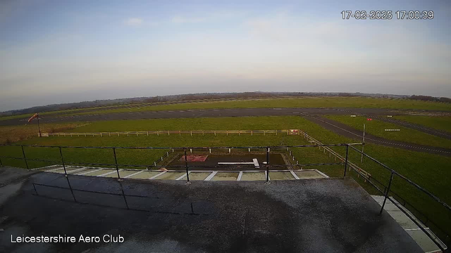 A view from a webcam at Leicestershire Aero Club showing a large green field and a runway. In the foreground, there is a railing along a ledge. The runway appears to be paved and has markings for takeoff and landing. A small area at the center has a white shape, likely a helicopter pad, surrounded by a gravel area. In the background, a clear sky is visible with a few clouds. The scene is calm and open, with no aircraft present.