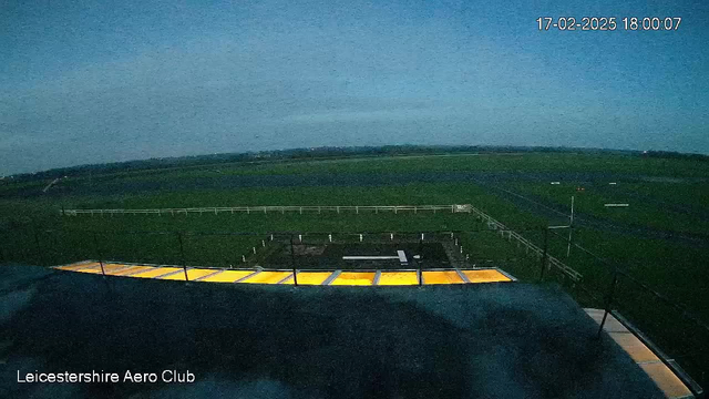 A view from a webcam at the Leicestershire Aero Club shows a landscape with an expansive green field. The foreground features a flat rooftop with yellow and gray surfaces. Below, there is a portion of the airstrip with markings, surrounded by white fence posts. The sky appears clear with a soft blue hue, suggesting it is late afternoon. The timestamp indicates the image was captured on February 17, 2025, at 18:00.