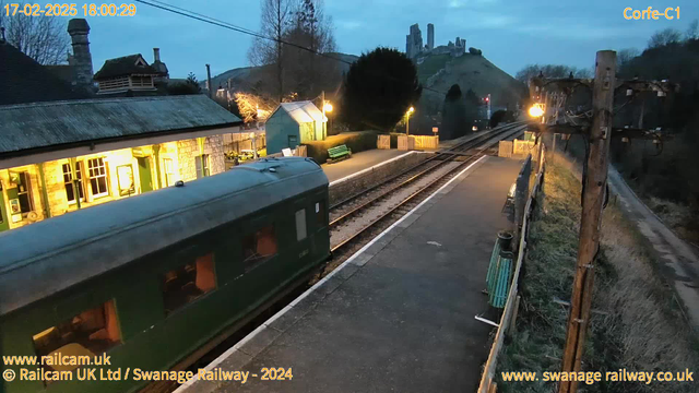 A train carriage, painted green, is positioned on a railway platform at dusk. The scene is set in Corfe, with a historic building featuring large windows and an illuminated interior on the left. There are several benches along the platform, and the landscape in the background includes a hill with ancient stone ruins. A warm glow from a nearby lamp enhances the atmosphere, while overhead power lines stretch across the sky. The image conveys a serene yet slightly nostalgic railway setting.