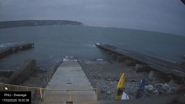 A coastal scene at twilight showing a rocky shoreline and a jetty extending into calm waters. The sky is overcast with a slight bluish tint. A set of stairs leads down to the water, to the left of which are two kayaks, one yellow and one blue, on a patch of gravel. The jetty has a railing and appears to be in the distance, with waves gently lapping against the structure. The background features cliffs partially shrouded in mist.