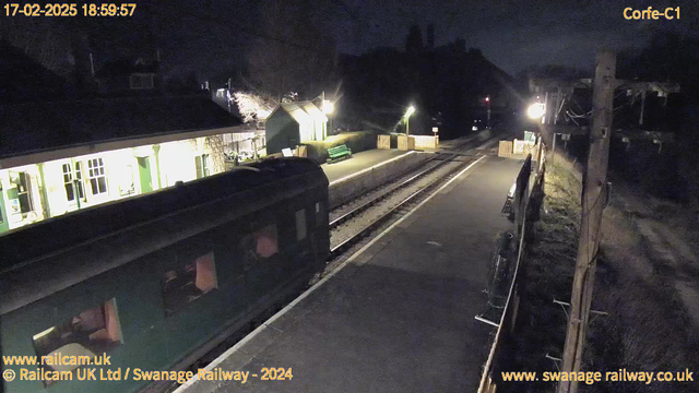 A dimly lit railway station at night features a green train on the left side of the image. The platform is empty, with a green bench and wooden fencing visible. Surrounding buildings are faintly illuminated, with some trees and a hint of a hillside in the background. Street lamps provide minimal lighting, casting soft shadows.