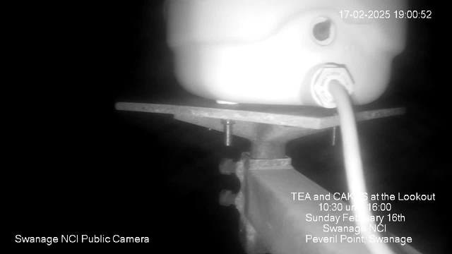 A black and white image from a webcam displaying the lower part of a camera mounted on a metal platform. The camera is positioned at a slight angle, showing a white rounded object above it. A wire is attached to the camera, connecting to a visible port. The background is mostly dark, and there is text at the bottom indicating it is from a public camera in Swanage NCI. Additional text presents information about a tea and cakes event, including the date and time, along with location details.