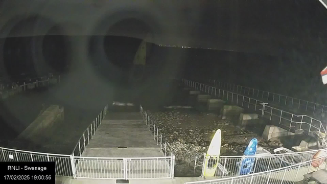 A dark scene shows a quay or marina area at night. A partially visible dock extends into the water on the left, lined with a white railing. Two colorful items resembling kayaks, one yellow and one blue, are positioned on the right side near the edge of the dock. The water appears still, with a rocky shoreline visible. Some indistinct shapes are seen further out in the water, and distant lights suggest other structures or land in the background.  The date and time displayed in the lower left corner indicate February 17, 2025, at 7:00 PM.