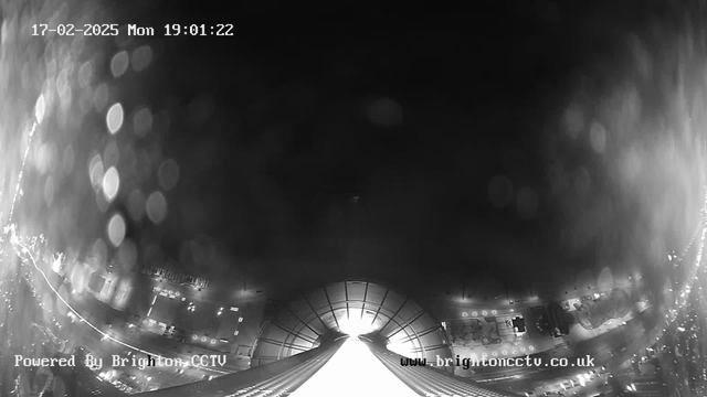 A black and white webcam image taken at night, with a timestamp showing 17th February 2025, 19:01:22. The image features bright light at the center, likely from a street or a building, radiating outwards. The edges contain blurred circular light reflections suggesting rain or condensation on the camera lens. On the bottom left, there is text that reads "Powered By Brighton CCTV," and the bottom right features the website address "www.brightoncctv.co.uk." The scene appears dim, with only the central light source providing visibility.