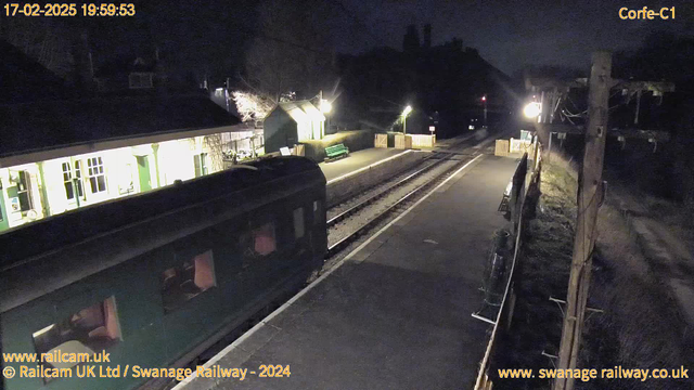 A dimly lit railway station scene at night shows a green train parked on the left side of the image. The platform is empty with a few wooden benches and a green bench positioned along the edge. In the background, there is a building with light coming from its windows, indicating activity inside. Street lamps illuminate parts of the platform, casting soft light. The railway tracks run parallel to the platform, leading off into the darkness on the right side of the image. There are some trees visible in the background, adding to the nighttime ambiance.