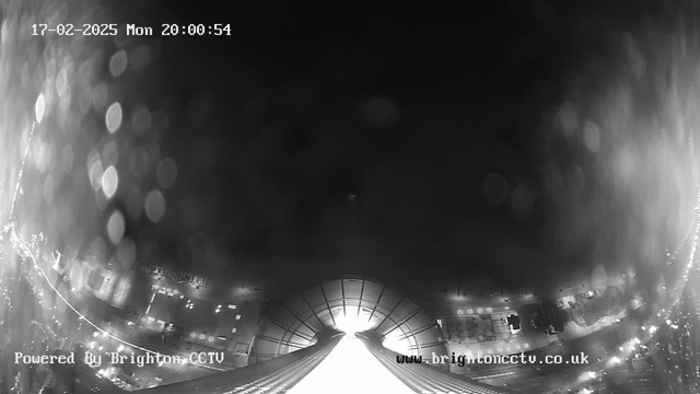 A black and white image from a webcam showing a view of a city at night. The scene is slightly blurry with light reflections, and the view appears to be focused on a structure leading into a brightly lit area in the center, resembling a tunnel or pathway. Numerous blurred lights suggest vehicles and street activity, while a timestamp indicates it is 20:00:54 on February 17, 2025. The bottom left corner includes a watermark indicating it is powered by Brighton CCTV.