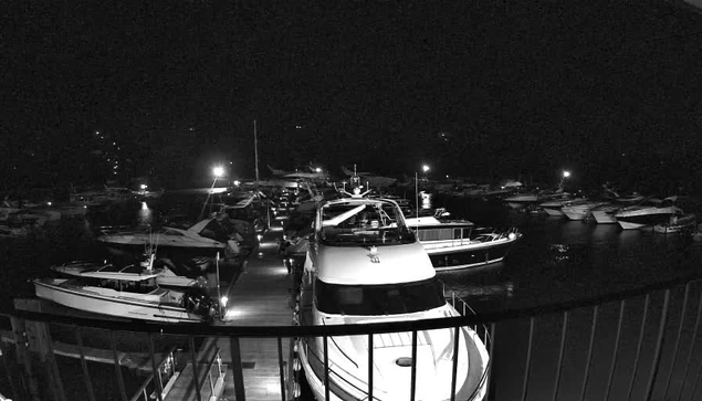 A nighttime view of a marina with several boats docked. The scene is illuminated by lights from the boats and the dock, creating reflections in the still water. A variety of boats of different sizes are visible, with a prominent white boat in the foreground. The background shows more boats and some distant lights, suggesting activity along the shore. The overall atmosphere is serene and calm, with dark surroundings contrasting with the illuminated areas.