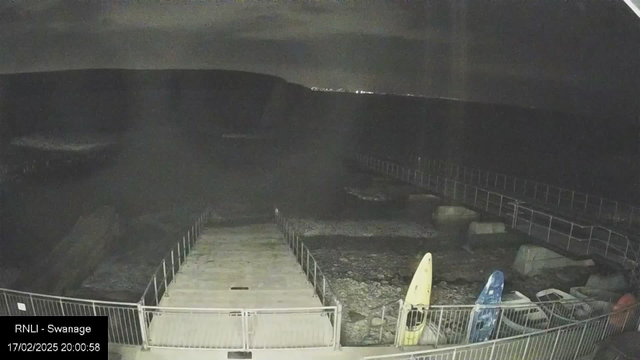 A dark scene shows a partially submerged dock leading out into a body of water, with rocky terrain visible on either side. Two kayaks, one yellow and one blue, are positioned on the right side of the dock. The area is illuminated by faint lights in the background, indicating some human presence. The sky is overcast, contributing to the overall dimness of the scene. A timestamp in the corner indicates the date and time of the image.