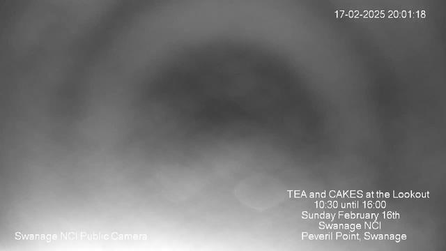 A blurred black and white webcam image showing indistinct shapes. In the lower right corner, there is text detailing an event: "TEA and CAKES at the Lookout, 10:30 until 16:00, Sunday February 16th, Swanage NCI, Peveril Point, Swanage." The current date and time are displayed in the upper right corner: "17-02-2025 20:01:18." The left corner includes "Swanage NCI Public Camera."