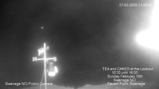 A dark and cloudy sky is depicted, with a faint light source visible. In the foreground, a weather vane with a flag is positioned vertically, marked with the letters "N" and "S" to indicate north and south. Text at the bottom announces an event for tea and cakes at the Lookout, providing details including the date, time, and location in Swanage. The overall image has a monochromatic appearance, primarily in shades of black and white.