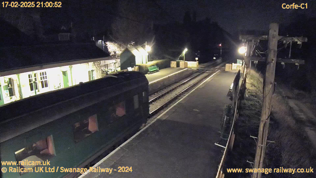 A dimly lit railway station at night. A green vintage train is parked on the left side of the image, with slightly illuminated windows suggesting occupancy. In the background, the platform features a wooden fence, a green bench, and a small shelter with a white roof. Lanterns along the platform cast a soft glow, while the surrounding area is dark, indicating a quiet and peaceful setting. The tracks are visible, leading off into the distance.