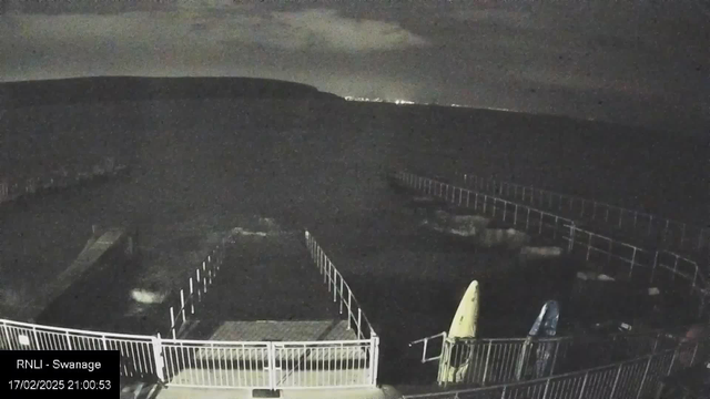 A dark nighttime scene shows a partially visible pier over water. The pier is surrounded by railings, with a few boats resting on the water. The sky has some clouds, and distant lights can be seen on the horizon. The time and date are displayed on the bottom left corner.