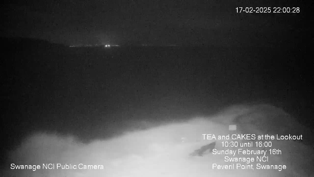 A dark, nighttime image showing a view of the sea with faint lights on the horizon. The foreground features blurred white shapes, possibly waves or sand. Text overlays indicate that this is a webcam image from Swanage NCI Public Camera, with event details for tea and cakes at the Lookout. The date and time show February 17, 2025, at 22:00:28.