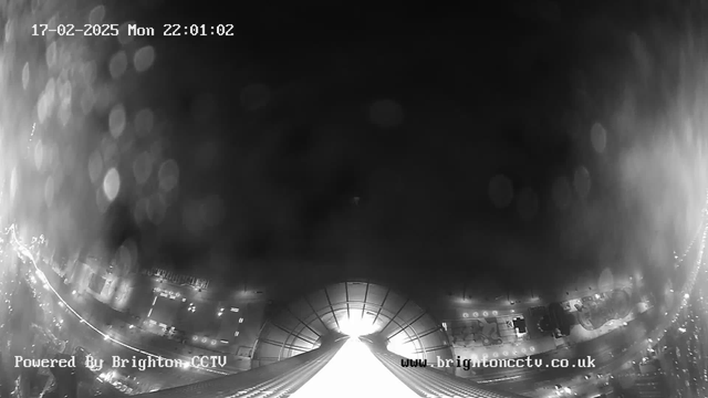 A black and white webcam image showing an urban landscape at night. The foreground features a bright pathway leading towards an open area, while faint circular light reflections are visible. In the background, there are blurred outlines of buildings and vehicles, with some lights indicating traffic. The timestamp shows it is Monday, February 17, 2025, at 22:01:02. The bottom of the image includes the text "Powered By Brighton CCTV" and a website URL.