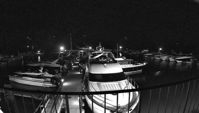 A nighttime scene of a marina with several boats moored along a wooden dock. The image is predominantly dark, with artificial lights illuminating some of the boats and reflecting off the water. The boats vary in size and type, with visible railings and features such as cabins and masts. The overall atmosphere is calm and serene, with a few bright spots contrasting against the darkness.