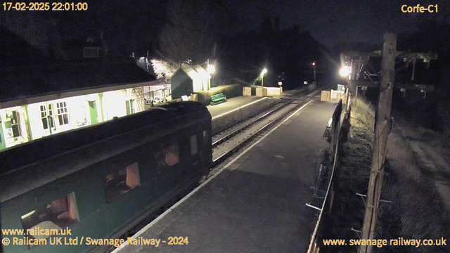 A dimly lit railway station at night. The platform is mostly empty, with a green bench visible on the left side. A dark green train car is partially in view on the left. The background features a building with lighted windows, and a wooden fence surrounds part of the area. There is a pole with a light on the right side illuminating part of the scene, and faintly visible train tracks stretch into the distance.