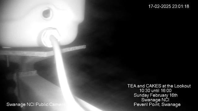 A dimly lit webcam image shows a white object partially visible, likely a camera or sensor, with a cable extending from it. The background is dark, obscuring details, but there is a horizontal surface below the object, possibly a table or platform. In the lower section of the image, text includes event information about "TEA and CAKES at the Lookout" on February 16th, along with the location "Swanage NCI" and "Peveril Point, Swanage."